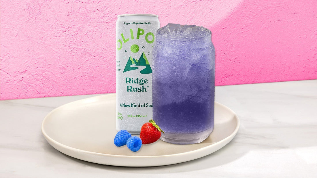 Purple drink in a glass with next to a OLIPOP Ridge Rush can on a white plate with fruit against a pink background