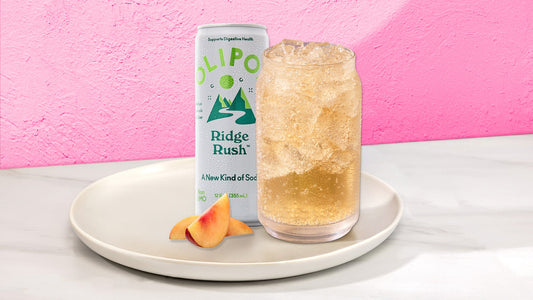 Peach drink in a glass with next to a OLIPOP Ridge Rush can on a white plate with fruit against a pink background