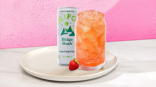 Orange drink in a glass with next to a OLIPOP Ridge Rush can on a white plate with fruit against a pink background
