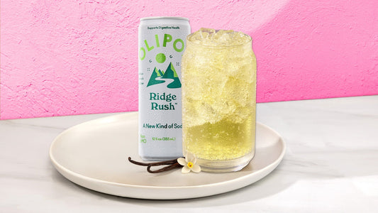 Light yellow drink in a glass with next to a OLIPOP Ridge Rush can on a white plate against a pink background