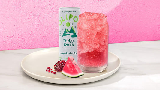 Pink drink in a glass with next to a OLIPOP Ridge Rush can on a white plate with fruit against a pink background